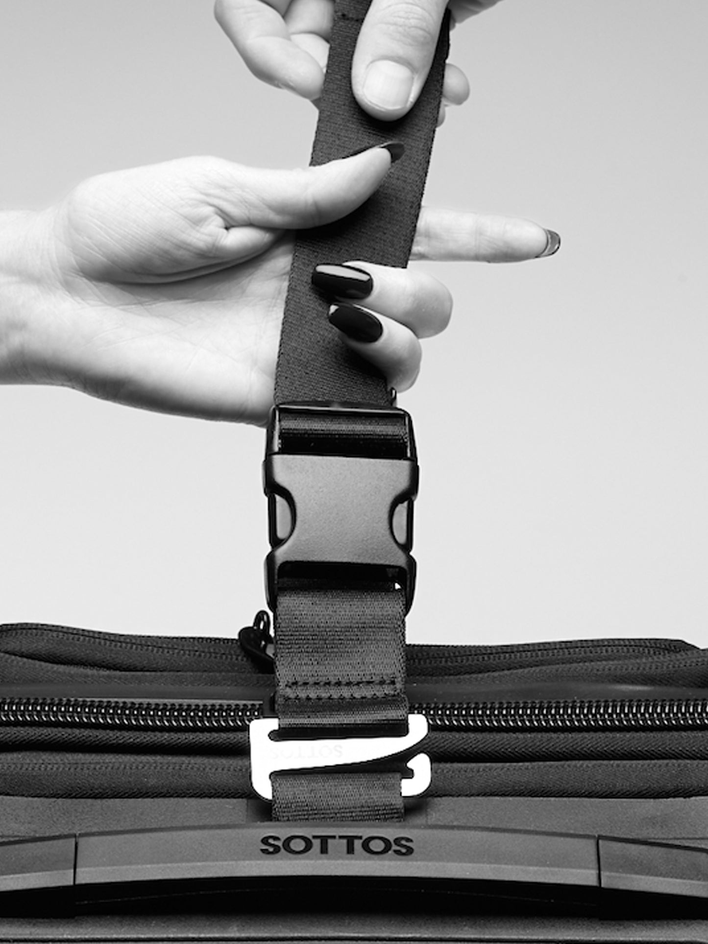 A black and white photo showing hands tightening a nylon strap on the top of the SOTTOS luggage bag.